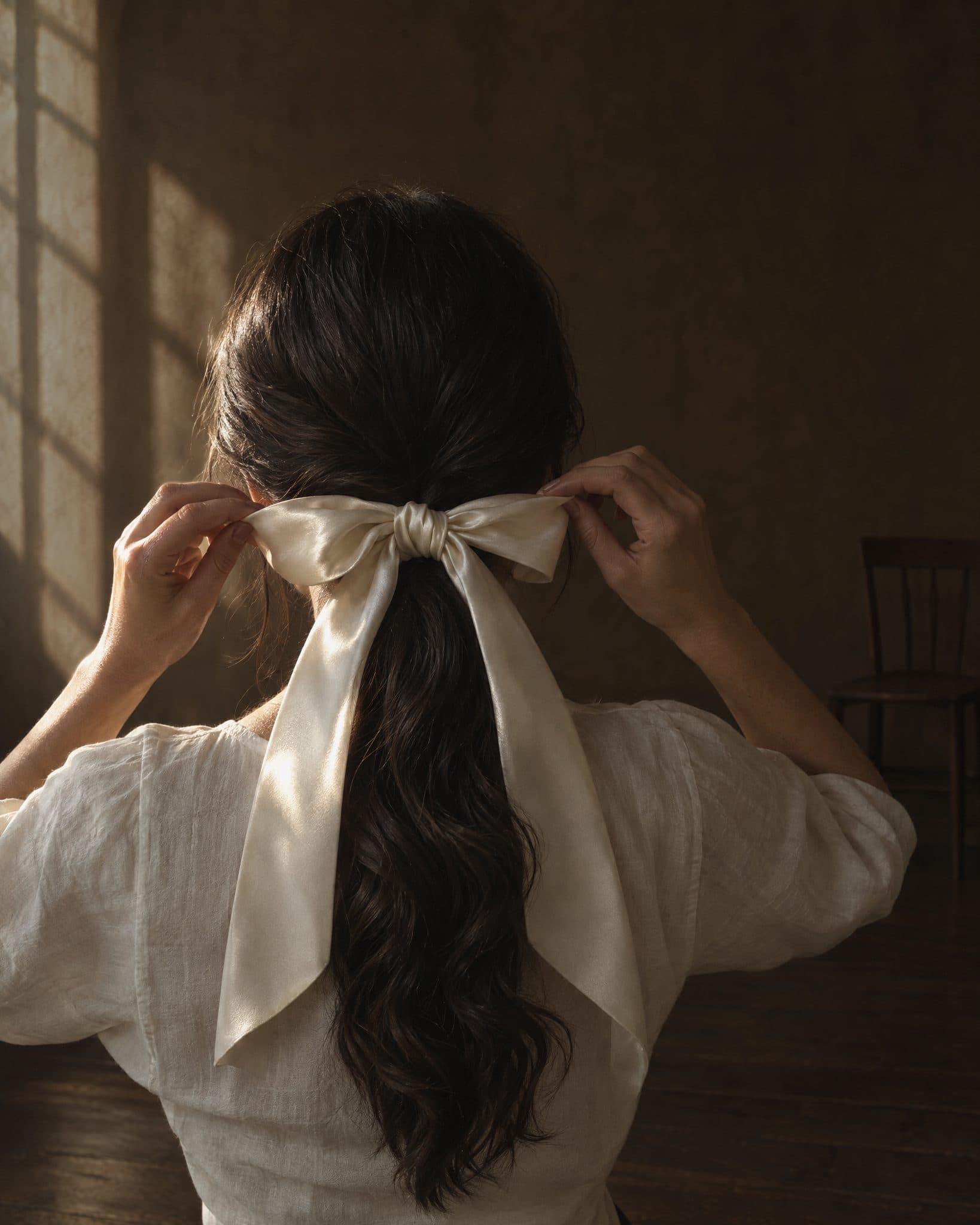 A woman seen from behind tying an ivory satin bow at the nape of her neck.